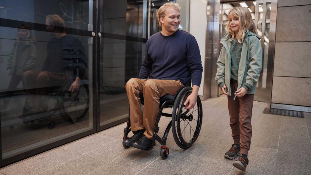 Un giovane uomo sorridente in sedia a rotelle insieme alla figlia piccola mentre si dirigono verso un ascensore accessibile in un edificio moderno.