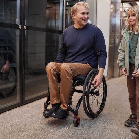 Un giovane uomo sorridente in sedia a rotelle insieme alla figlia piccola mentre si dirigono verso un ascensore accessibile in un edificio moderno.